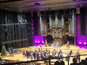 Scena filharmonii z muzykami i instrumentami oraz stojącą udekorowaną choinką po lewej stronie