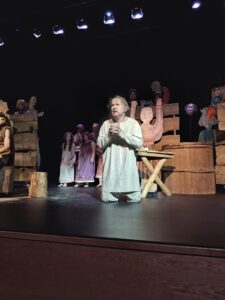 Scena z postacią w płóciennym stroju klęczącą na tle drewnianej scenografii