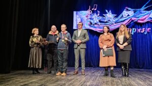 Sześć dorosłych osób stojących na drewnianej scenie z niebieską kotarą w tle