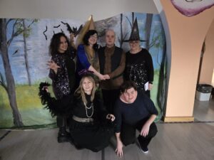 Sześcioro dorosłych w czarnych strojach stojących w grupie na tle dekoracji przedstawiającej łąkę z drzewami