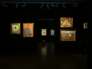 Wiszące w ciemnej przestrzeni siedem kolorowych obrazów olejnych przedstawiających ludzi