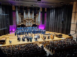 Wnętrze filharmonii z widzami i sceną z muzykami z instrumentami oraz chórem w niebieskich strojach stojącym na drugim planie