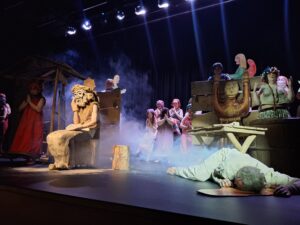 Zdjęcie sceny ze spektaklu przedstawiające rzeźbę Chrystusa Frasobliwego i aktorów na tle drewnianej scenografii