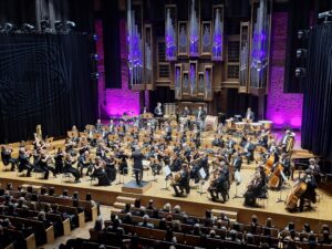 Drewniana scena w filharmonii z siedzącą na niej orkiestrą z instrumentami i dyrygentem oraz w tle na ścianie organy oświetlone fioletowym światłem