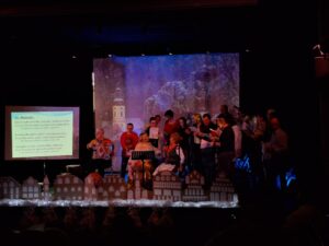 Duża grupa osób ze śpiewnikami stojących na scenie oświetlonej na niebiesko i otoczonych scenografią w formie ośnieżonych domków