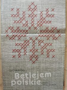 Fragment ulotki informacyjnej o spektaklu Betlejem polskie z wyszytym krzyżykowo białymi i czerwonymi nićmi płatkiem śniegu na szarym płótnie
