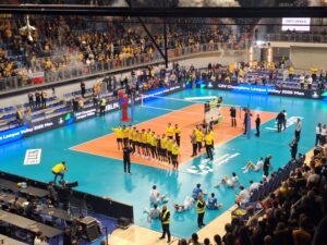 Hala sportowa wypełniona kibicami i drużyną pozującą do fotografii na linii końcowej boiska do siatkówki