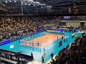 Hala sportowa wypełniona kibicami oraz dwoma drużynami stojącymi na liniach końcowej i początkowej boiska siatkowego