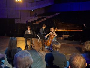 Na drewnianej scenie filharmonii trzy siedzące osoby z instrumentami - mężczyzna z klarnetem mężczyzna przy fortepianie i kobieta z wiolonczelą