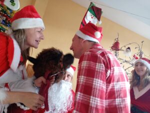 We wnętrzu dwie dorosłe osoby w mikołajowych przebraniach pochylone nad alpaką która stoi między nimi