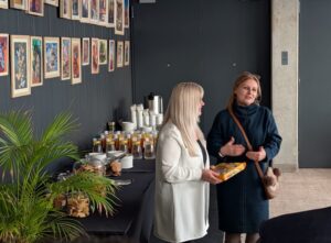 Dwie kobiety stojące przy kolorowych pracach plastycznych eksponowanych na ciemnoszarej ścianie