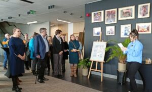 Grupa dorosłych osób stojąca przy pracach plastycznych eksponowanych na ciemnoszarej ścianie