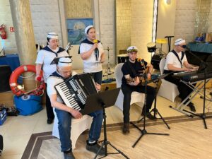 Grupa pięciu siedzących osób z instrumentami muzycznymi i z czapeczkami marynarskimi na głowach