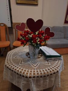 Trzy papierowe czerwone serca umieszczone w bukiecie czerwonych róż w szklanym flakonie stojącym na siedmiokątnym stole nakrytym ręcznie robioną białą serwetą