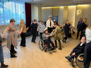 We wnętrzu grupa dorosłych osób tańczących w kółku oraz kilka osób tańczących w parach