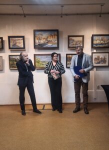 We wnętrzu trzy osoby dorosłe stojące na tle zawieszonych obrazów z pejzażami zimowymi i wiosennymi