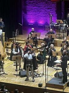 Widoczna część grupy członków orkiestry z instrumentami na scenie oraz stojący mężczyzna w stroju góralskim grający na trombicie