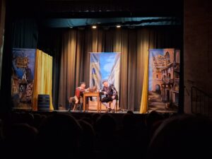 Na scenie dwóch mężczyzn w renesansowych strojach siedzących przy stole na tle dekoracji imitujących ściany kamienic