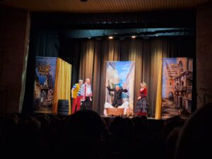 Na scenie kilka stojących osób w mieszczańskich strojach z których jedna rozrzuca białe koszule z wiklinowego kosza