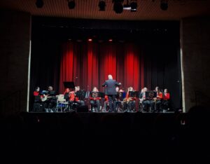 Na scenie z czerwoną kurtyną w tle kilkanaście siedzących osób z instrumentami dętymi oraz dyrygent