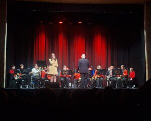 Na scenie z czerwoną kurtyną w tle kilkanaście siedzących osób z instrumentami dętymi oraz wokalistka i dyrygent