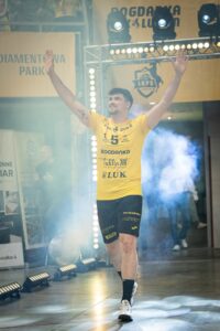 Uśmiechnięty mężczyzna z uniesionymi rękami w żółto-czarnym stroju wchodzący na sportową halę