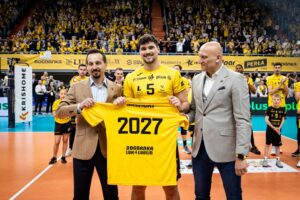 Zawodnik siatkarski w żółtej koszulce i po jego bokach pojedynczy mężczyźni w marynarkach stojący na boisku do piłki siatkowej w hali sportowej wypełnionej widzami