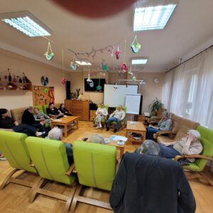 Grupa dorosłych osób siedząca we wnętrzu na zielonych fotelach ustawionych na planie kwadratu