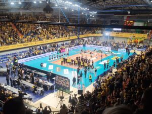 Hala sportowa wypełniona kibicami i drużyna pozująca do fotografii na boisku do siatkówki po zakończonym meczu