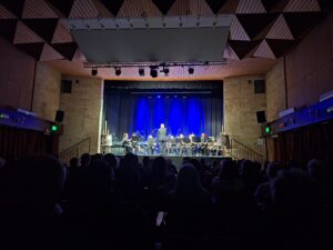 Oświetlona na granatowo scena widowiskowa z widocznymi muzykami z instrumentami oraz dyrygentem