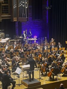 Scena filharmonii wypełniona muzykami trzymającymi instrumenty i siedzącymi przed pulpitami nutowymi i stojącym przed nimi dyrygentem