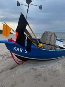 Częściowy widok niebieskiej łodzi z kolorowymi flagami i napisem Kar-5 leżącej na piaszczystej plaży oraz morze i chmurne niebo w tle