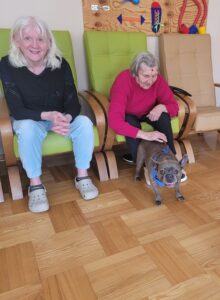 Dwie dorosłe osoby siedzące w dużych fotelach a jedna z nich jest pochylona do przodu i głaszcze siedzącego przed nią buldoga