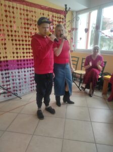 Dwie osoby w czerwonych ubraniach stojące obok siebie we wnętrzu i trzymające mikrofony w dłoniach