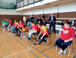 Grupa dorosłych osób siedząca na tapicerowanych krzesłach ustawionych przy bocznej linii boiska w hali sportowej