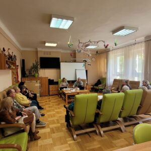 Grupa dorosłych osób siedzących w zielonych fotelach w sali o charakterze klubowym