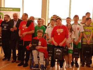 Grupa kilkunastu dorosłych osób z medalami zawieszonymi na szyjach stojąca na tle dużych okien sali gimnastycznej