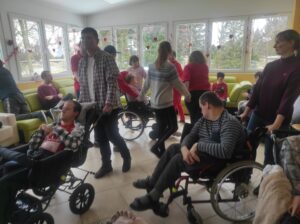 Grupa stojących osób we wnętrzu ubranych na czerwono oraz grupa osób na wózkach inwalidzkich