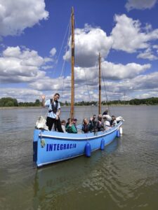 Płynąca niebieska dwumasztowa łódź z pasażerami na tle nieba z dużymi białymi chmurami
