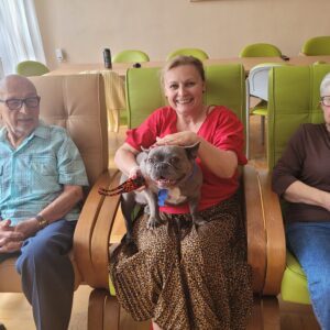Trzy dorosłe osoby siedzące w dużych fotelach a jedna z nich trzyma i głaszcze buldoga na swoich kolanach