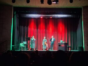 Trzy osoby stojące w otoczeniu instrumentów na tle czerwonej kutyny w sali widowiskowej