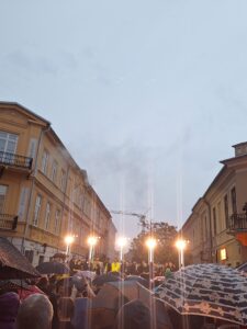 W perspektywie ulicy z kamienicami na pierwszym planie widoczni ludzie z parasolkami a nad nimi zawodnicy sportowi w żółtych oraz czarnych strojach stojący na tle pięciu reflektorów