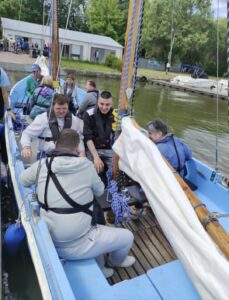 Widziana z góry grupa ośmiu osób siedzących na dwumasztowej niebieskiej łodzi