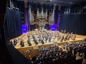 Wnętrze filharmonii z muzykami orkiestry na scenie oraz widzami siedzącymi w fotelach