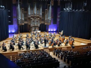 Wnętrze filharmonii ze sceną wypełnioną muzykami z instrumentami dyrygentem oraz publicznością siedzącą w fotelach