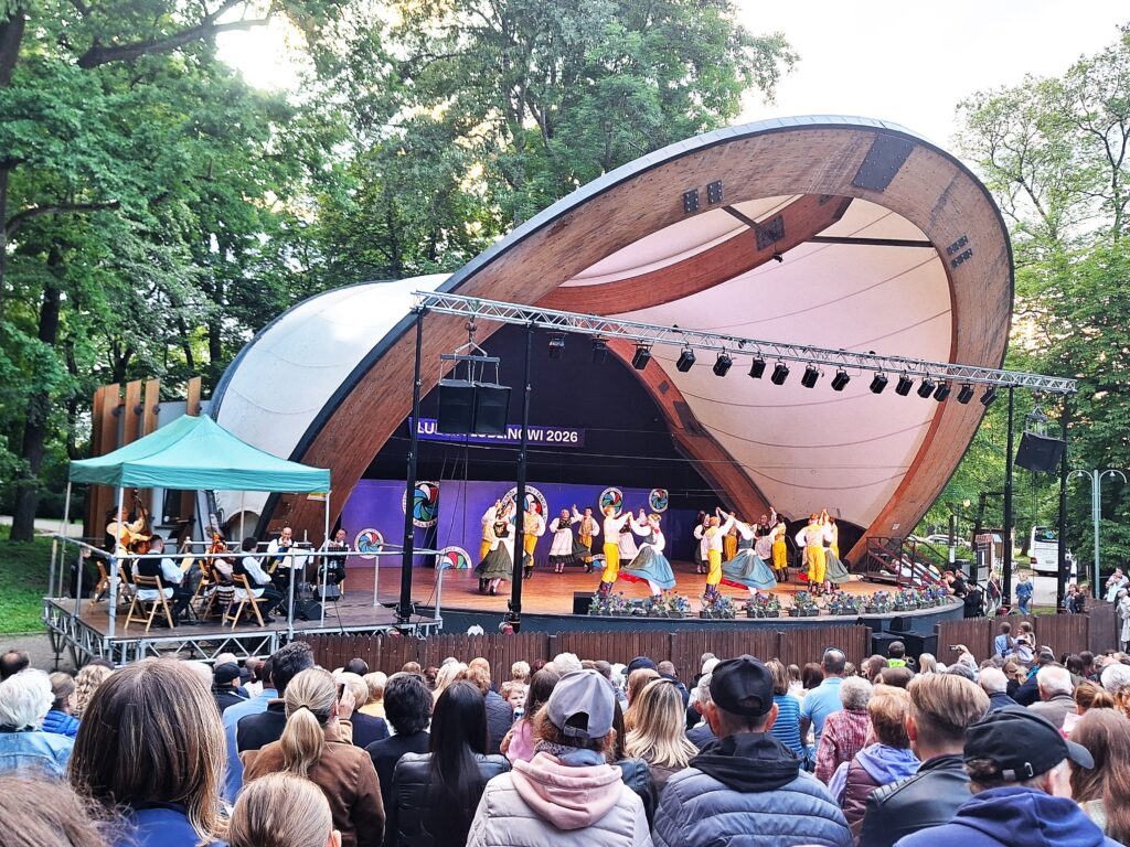Grupa par tanecznych w biało-żółto-czarnych strojach ludowych tańczących na scenie w muszli koncertowej