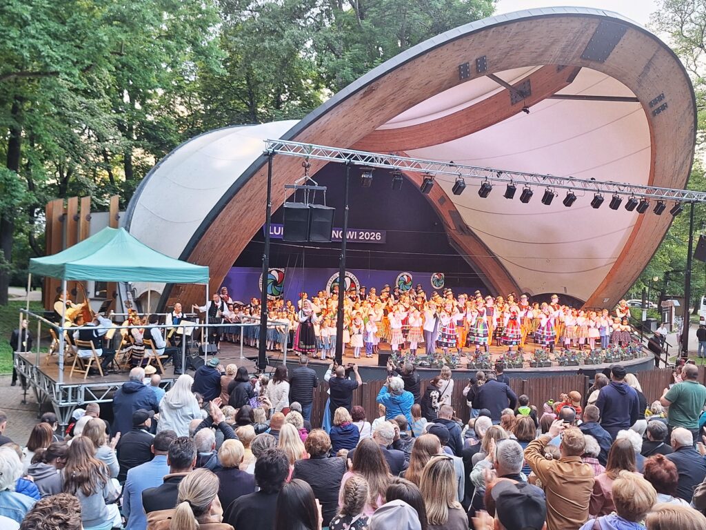 Bardzo duża grupa tancerzy w strojach ludowych stojąca na scenie w muszli koncertowej