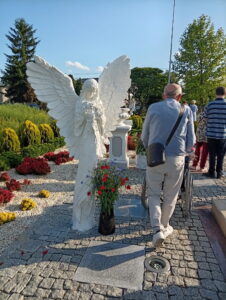 Biała rzeźba kobiecej postaci ze skrzydłami trzymającej kwiaty ustawiona na brukowanym placu