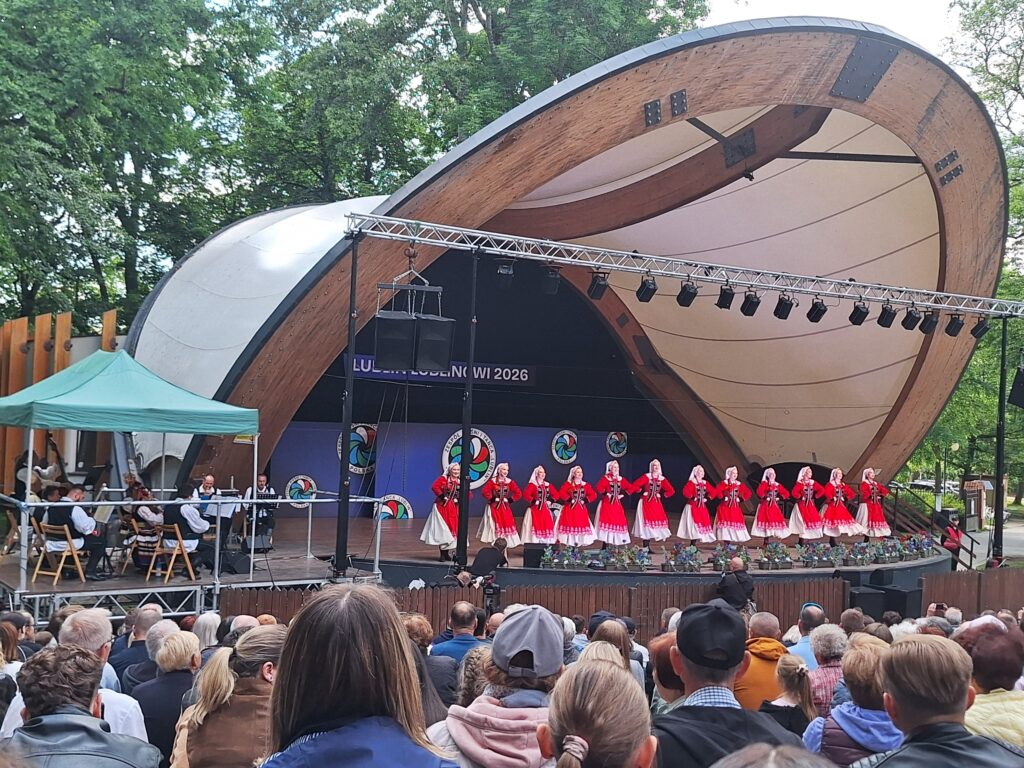 Grupa kobiet w czerwono-białych strojach ludowych tańcząca na scenie w muszli koncertowej