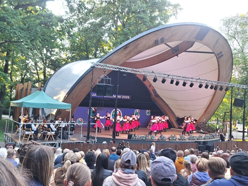 Grupa par tanecznych w różowo-białych strojach ludowych tańczących na scenie w muszli koncertowej